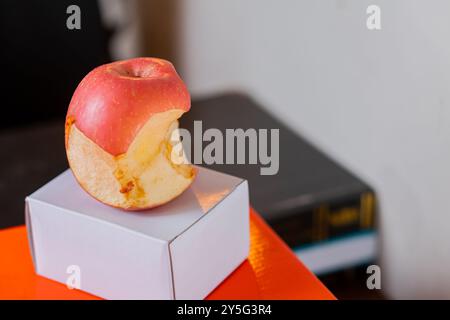 Ein teilweise gegessen roter Apfel, der auf einer kleinen weißen Box liegt. Der Apfel wird mit einem Biss herausgenommen, der sein saftiges Inneres offenbart. Der Hintergrund ist unscharf Stockfoto