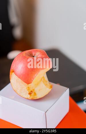 Ein gebissener roter Apfel, der auf einer kleinen weißen Box liegt, mit einem bunten Hintergrund. Der Apfel hat eine glänzende Oberfläche und einen teilweise freiliegenden Innenraum, Showcasin Stockfoto