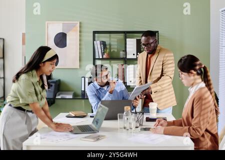 Das Büroteam diskutiert die Geschäftsstrategie mit Schwerpunkt auf dem Tablet, das vom Teammitglied Standing gehalten wird. Papiere und Laptops liegen auf dem Tisch, während verschiedene Teams sich mit Diskussionen beschäftigen Stockfoto