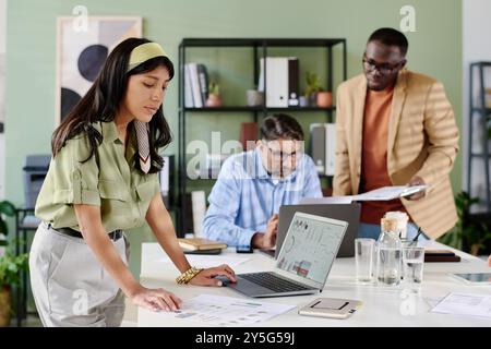 Gruppe von Arbeitnehmern, die am Tisch mit Dokumenten und Laptops zusammenarbeiten, konzentriert sich auf die Projektbesprechung. Büroumgebung mit Regalen und Grünflächen im Hintergrund Stockfoto