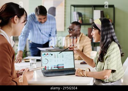 Multiethnisches Team diskutiert Geschäftsanalysen auf Laptops, die am Konferenztisch sitzen, Ideen austauschen und Daten analysieren. Professionelles Ambiente mit offener Atmosphäre und modernem Bürodesign Stockfoto