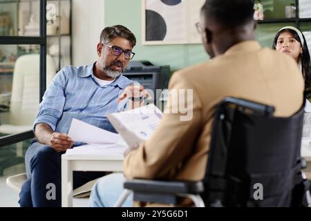 Verschiedene Fachleute diskutieren Dokumente in einer modernen Büroumgebung, wobei Kaukasier das Gespräch führen und Afroamerikaner sich Notizen machen Stockfoto
