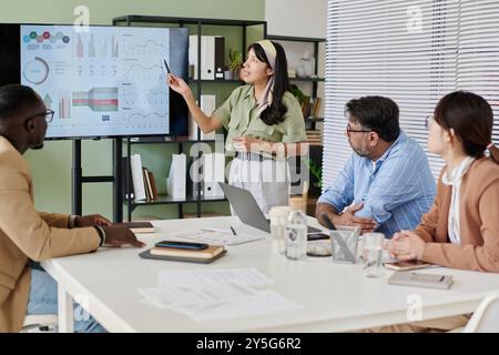 Gruppe von Kollegen, die Geschäftsdaten im Besprechungsraum diskutieren, mit Diagrammen auf dem Bildschirm und verschiedenen Dokumenten auf dem Tisch. Präsentator erklärt Diagramme, während andere aufmerksam zuhören Stockfoto