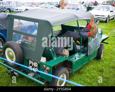 MINI MOKE MIT EINEM MANN, DER ENTSPANNT EIN BUCH AUF EINEM TABLET MIT STIEFELN AUF EINEM KLASSISCHEN SAINT MAWGAN DAMPFRALLYE LIEST Stockfoto