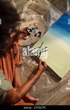 Person, die lebendige Landschaftsmalerei auf Leinwand erstellt, die auf dem Boden platziert ist, mit einer Palette, um Farben zu mischen und sie mit einem Pinsel aufzutragen. Glatter, mit sanften Strichen bemalter Himmel Stockfoto