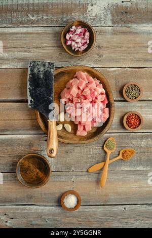 Frisch gehacktes Fleisch wird auf einem Holzteller zusammen mit verschiedenen Gewürzen, Knoblauch und Schneideutensilien auf einem rustikalen Holztisch platziert Stockfoto