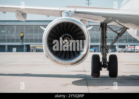 Maschinenhaus des Flugzeugmotors. Vorderansicht des Turbofan-Triebwerks unter einem Flügel eines Passagierflugzeugs in der Nähe des Fahrwerks. Flughafenterminal im Hintergrund. Stockfoto