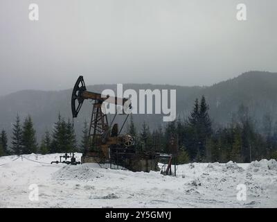 Ölpumpe. Anlagen für die Öl- und Gasindustrie. Ölfeld-Pumpenheber und Ölraffinerie im Winter mit Schnee, Bergen und Wald im Hintergrund. Im Schnee Stockfoto