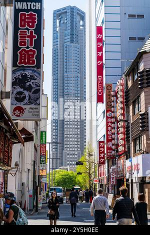 Blick auf Chuo dori, eine enge Einkaufsstraße in Shinjuku mit dem riesigen Tokyo Metropolitan Gebäude am Ende, das den Himmel an einem sonnigen Tag erfüllt. Stockfoto