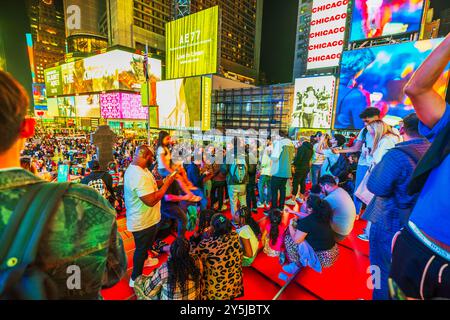 Die Menschen versammelten sich nachts auf der roten Treppe am Times Square, umgeben von hellen Plakatwänden und Menschenmassen in New York City. USA. Stockfoto