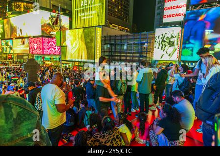 Leute sitzen und stehen nachts auf der roten Treppe am Times Square, umgeben von hellen Plakatwänden und Menschenmassen in New York City. USA. Stockfoto