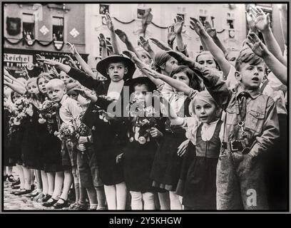ANSCHLUSS AUSTRIAN CHILDREN HEIL HITLER SALUTE 1930er NS-Propagandabild österreichischer Kinder, darunter ein Hitler-Jugendjunge in Uniform, der Adolf Hitler mit traditionellem Heil Hitler-Salut mit erhobenen Armen begrüßte. Hakenkreuz-Symbol an der Wand im Hintergrund. Wien (Hinweis: Jüdisches Eigentum und später „erworben“ von den Nazis, REICHEL Textilgeschäft in b/g) Wien Österreich Stockfoto