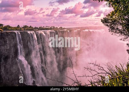 Atemberaubende Victoria Falls, Wasserfall, der unter einem wunderschönen, ruhigen Himmel bei Sonnenuntergang über felsigen Klippen stürzt und einen Nebel erzeugt, der zwischen üppigen Bäumen aufsteigt Stockfoto