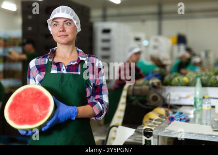 Arbeiterin posiert in der Gemüsefabrik mit der neuen Ernte der organischen Wassermelonen, hält saftige Wassermelone in der Mitte geschnitten Stockfoto