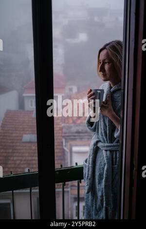 Am frühen Morgen steht eine Frau in einem Gewand still auf ihrem Balkon, hält eine dampfende Tasse Tee und blickt in den nebelbedeckten Straßenbelag Stockfoto