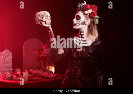 Frau posiert mit Schädel in der Hand, spielt Hamlet-Szene, während sie Skelett-Make-up trägt. Makabre Dame, gekleidet als La Catrina Charakter am Tag des Dead Events im Studio Hintergrund mit rotem Licht Stockfoto