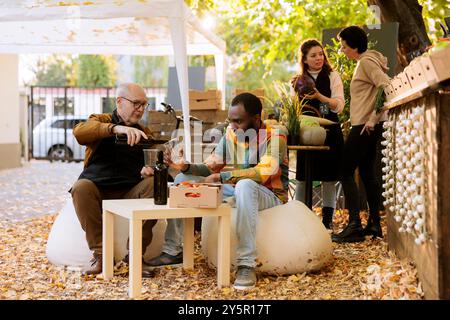 Multikulturelle männliche Individuen genießen ein Glas Naturwein an einem Tisch neben einem Bauernmarkt. Verschiedene Kunden besuchen ein Erntefest und probieren frisch geerntete Produkte. Stockfoto