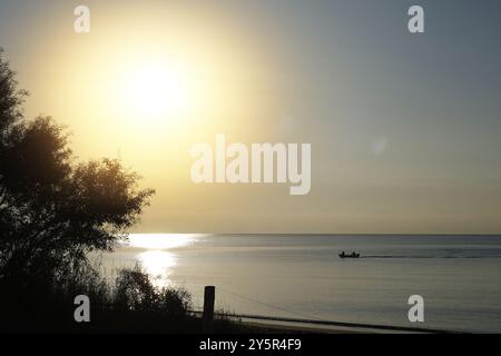Ein kleines Boot mit zwei Personen gleitet während eines hellen Sonnenaufgangs auf ruhigem Wasser, mit einem goldenen Leuchten, das auf der Meeresoberfläche reflektiert. Stockfoto