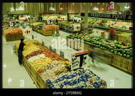 In den 1980er Jahren stöbern die Käufer in einem großen Supermarkt, der sich auf frisches Obst und Gemüse wie Wassermelonen, Zwiebeln und Kartoffeln konzentriert. Die hellen Schilder des Stores fördern sein Engagement für Qualität und Frische und laden Kunden in die Bereiche „Gourmet Kitchen“ und „Frische First“ ein. Stockfoto