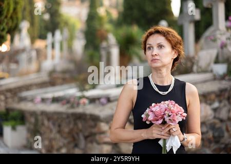 Eine verärgerte Frau in schwarzem Kleid, die einen geliebten Menschen verloren hat, bringt Blumen ins Grab Stockfoto