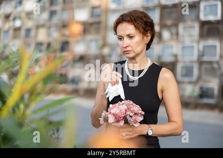 Eine verärgerte Frau in schwarzem Kleid, die einen geliebten Menschen verloren hat, bringt Blumen ins Grab Stockfoto