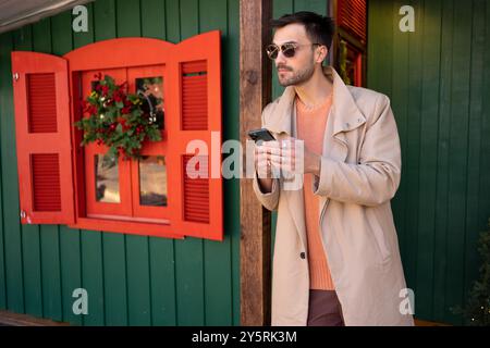Nachdenklicher Mann in beigem Trenchcoat, der vor dem Treibhaus ein Smartphone hält. Ein hübscher junger Mann schaut weg, während er tagsüber am roten Fenster steht. Stockfoto