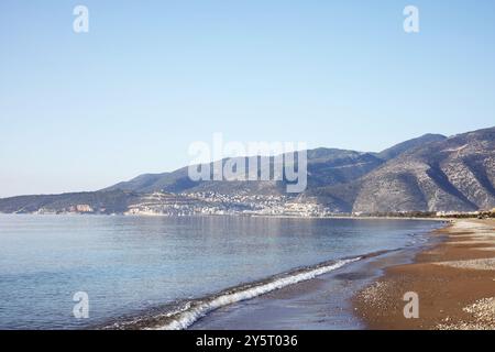 Ein ruhiger mediterraner Strand mit sanften Wellen über die Küste. Die fernen Berge umrahmen die Küste, mit einem klaren blauen Himmel darüber. Stockfoto