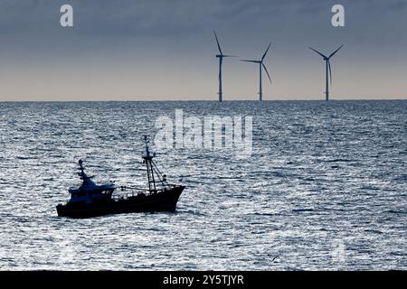 Ein Fischerboot fährt über den Ozean, umgeben von Windturbinen in der Ferne. Die Szene fängt das ruhige Wasser in der Abenddämmerung ein Stockfoto