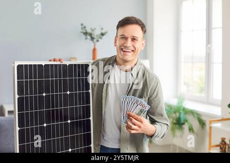 Glücklicher Mann, der ein Solarmodul hält und Geld zeigt, das er gespart hat, während er es benutzt Stockfoto