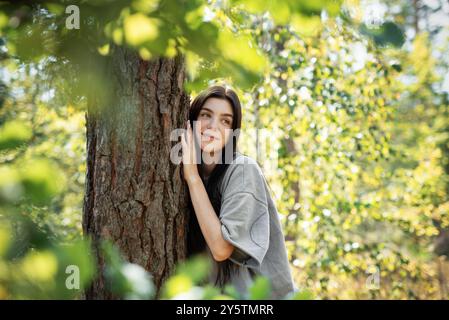 Eine Frau lehnt sich an einen Baum, blickt mit einem ruhigen Ausdruck nach oben, umgeben von leuchtend grünen Blättern in einem sonnendurchfluteten Wald. Stockfoto