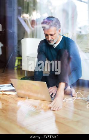 Homeoffice für einen schönen Mann. Mann, der auf dem Boden sitzt und am Laptop arbeitet. Durchgangsfenster. Telearbeit. Stockfoto