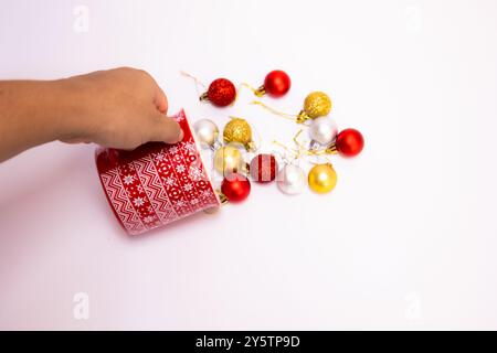 Weibliche Hand mit rotem Weihnachtsbecher und verstreuten goldenen, roten und silbernen Weihnachtskugeln auf weißem Hintergrund mit Blick von oben Stockfoto