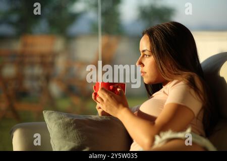 Profil einer ernsten Frau, die Kaffee trinkt und zu Hause bei Sonnenuntergang durch ein Fenster schaut Stockfoto