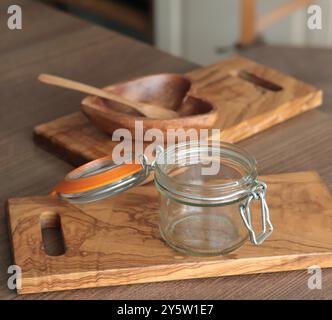 Leerer Glasbehälter auf Olivenholzbrett und herzförmige Marmeladeschale aus Holz. Küchenwerkzeuge für die Zubereitung von Obst. Stockfoto