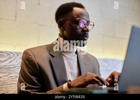 Geschäftsmann mit Brille, der am Laptop arbeitet, während er am Arbeitsplatz mit hölzernem Hintergrund sitzt, sich auf den Bildschirm konzentriert und über den Arbeitsprozess nachdenkt Stockfoto