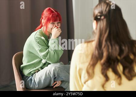 Frau mit roten Haaren, grüne Pullover und Jeans, lächelt, während sie auf dem Stuhl sitzt. Verschwommene Person mit langen Haaren im Vordergrund, wodurch der Fokus auf die rothaarige Frau gelegt wird Stockfoto
