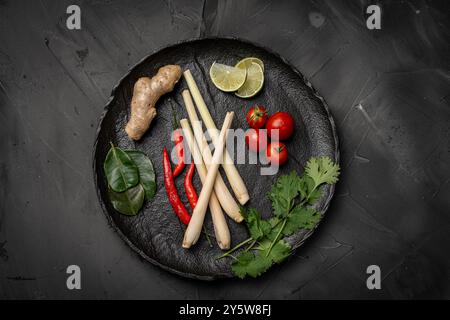 Zutaten für die Herstellung von Tom Yum Kung. Zitronengras, Chili-Pfeffer, Ingwer, Galgant, Limette, Koriander und Kirschtomaten auf einem schwarzen Teller vor einem dunklen Ba Stockfoto