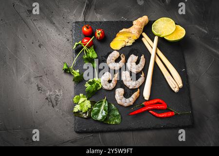 Zutaten für die Herstellung von Tom Yum Kung. Zitronengras, Chili-Pfeffer, Ingwer, Galgant, Limette, Koriander, Kirschtomaten und rohe Garnelen wieder auf einem schwarzen Teller Stockfoto