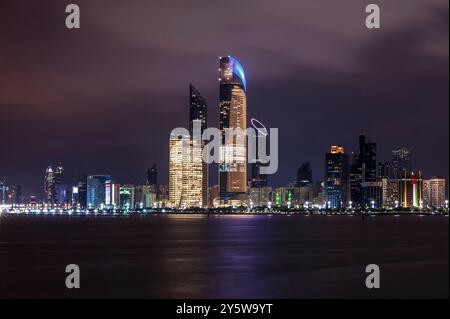 Nachtblick auf Abu Dhabi Corniche. Helle Skyline mit farbenfrohen Lichtern, die sich auf ruhigem Wasser spiegeln. Hochhäuser wie Burj Mohammed bin Rashid Stockfoto