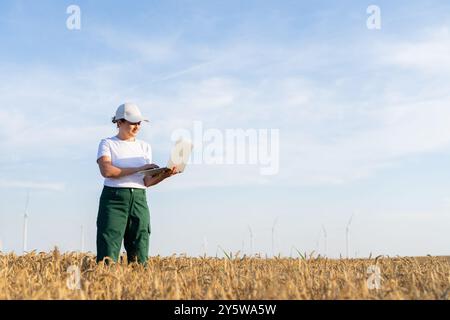 Farmerin mit weißer Mütze und T-Shirt mit Laptop auf dem landwirtschaftlichen Feld bei Sonnenuntergang. Windräder im Hintergrund... Stockfoto