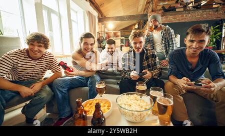 Gruppe von Freunden mit Controllern, Augen auf dem Bildschirm gesperrt. Snacks und Getränke tanken die Intensität, während sie anfeuern und im hitzigen virtuellen Spiel antreten. Stockfoto