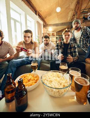 Unbeschwerte Atmosphäre mit Freunden, die zusammen spielen, Augen auf dem Bildschirm, Lachen durchbricht die Spannung des Spiels. Snacks und Getränke machen es klassisch. Stockfoto