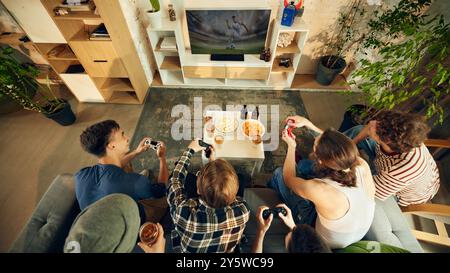 Blick von oben auf Freunde, die sich um den Tisch gedrängt haben, die Controller fest greifen, Snacks in Reichweite, während sie auf der Großbildleinwand kämpfen. Stockfoto