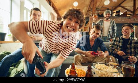 Ein Spieler lehnt sich nah an und hat ein großes Grinsen, während er den Controller packt. Die Energie der Gruppe ist hoch, Snacks und Getränke sind auf dem Tisch verteilt. Stockfoto