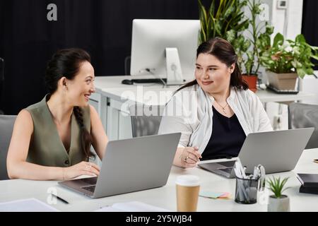 Zwei professionelle Frauen führen ein lebhaftes Gespräch, während sie im Büro an ihren Laptops arbeiten. Stockfoto