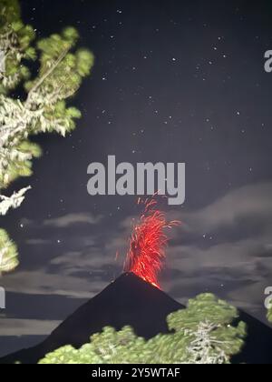 Nächtlicher Blick auf einen ausbrechenden Vulkan mit Lava und Sternen am Himmel, eingerahmt von Bäumen. Stockfoto