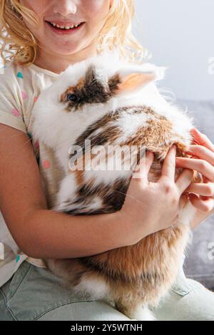 Ein junges Mädchen lächelt hell, während es einen flauschigen Häschen nahe an ihre Brust hält. Das sanfte Sonnenlicht hebt das Pelz des Kaninchens hervor und sorgt für ein warmes und liebevolles Gefühl Stockfoto