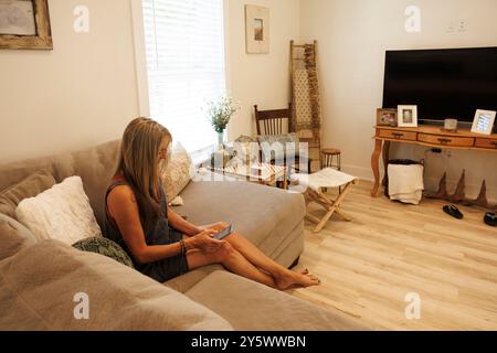 Frau, die mit einem Smartphone auf einer Couch sitzt, in einem gemütlichen Wohnzimmer mit stilvollem Dekor und einem großen Fernseher, Florida, USA Stockfoto