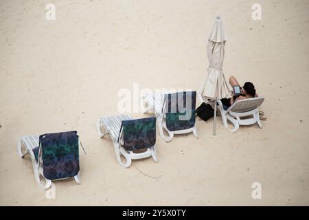 Aus der Vogelperspektive auf einen Sandstrand mit einer Person, die sich auf einer Liege unter einem geschlossenen Sonnenschirm entspannt, flankiert von leeren Stühlen und Handtüchern, Titikaveka, Rarotonga, Cook Islands Stockfoto