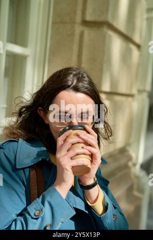 Junge Erwachsene Frau in blauem Mantel, die an einem sonnigen Tag Kaffee aus einer Tasse zum Mitnehmen schlürft. Stockfoto
